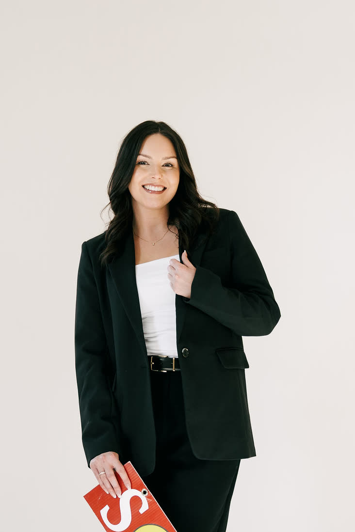 Samantha Johnson smiling, one hand at the lapel of her black suit, holding a red SOLD sign.