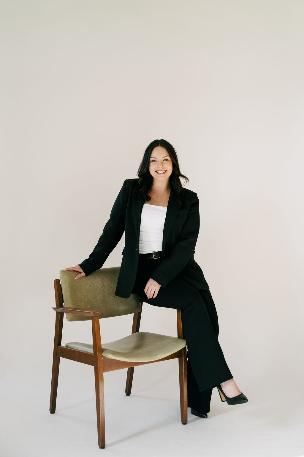 Samantha Johnson smiling, seated on the back of a wooden chair, wearing a black suit.
